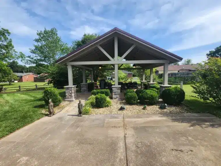 Large outdoor pavilion with white timber frame, stone column bases, and metal roof surrounded by shrubs and lawn.