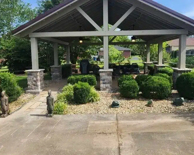 Outdoor pavilion with stone columns, ceiling fans, and patio seating, surrounded by trimmed shrubs and rock landscaping.