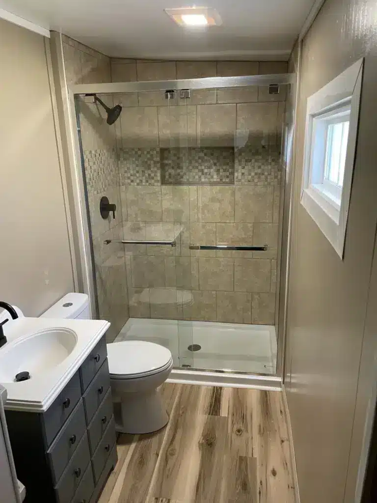 Small bathroom with tiled walk-in shower, glass sliding door, white toilet, gray vanity, and wood-look flooring.