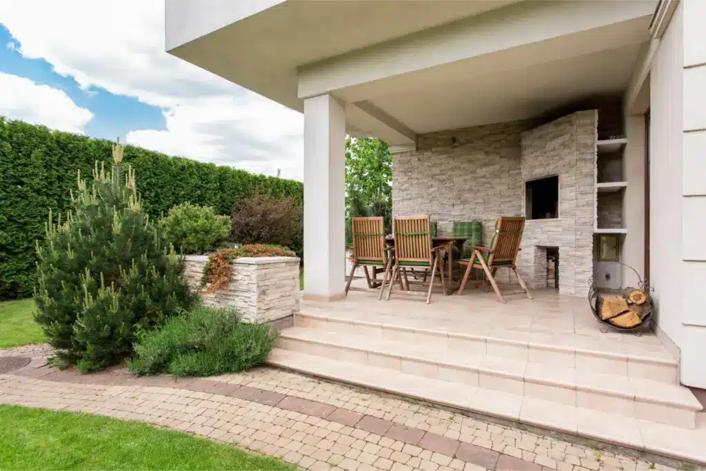 Covered tiled patio with wooden dining set, stone veneer fireplace, paver walkway, and landscaped garden with evergreen shrubs.