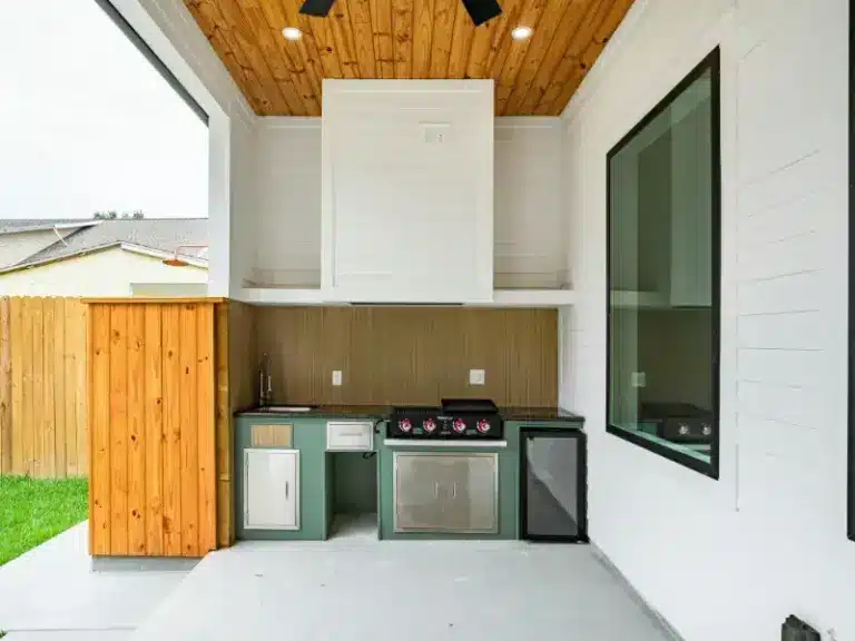 Covered patio with built-in outdoor kitchen featuring a gas grill, sink, mini fridge, and stained wood plank ceiling.