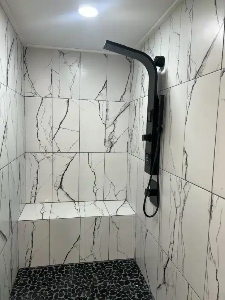 Walk-in shower with white marble-look wall tile, built-in bench, black pebble floor, and matte black shower panel.