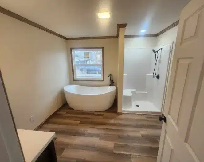 Bathroom with white freestanding soaking tub under a window, walk-in shower with black fixtures, and wood-look flooring.