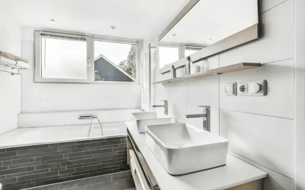Modern bathroom with double vessel sinks, wall mirror, built-in bathtub with gray tile surround, and a window.