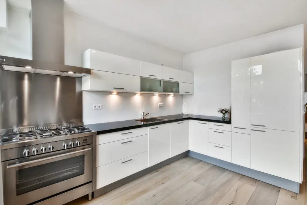 Modern kitchen with white gloss cabinets, black countertops, stainless steel range and hood, and light wood flooring.