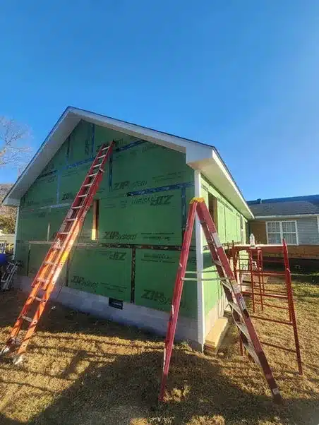 House under construction with green ZIP System wall sheathing, two extension ladders, and scaffolding along one side.