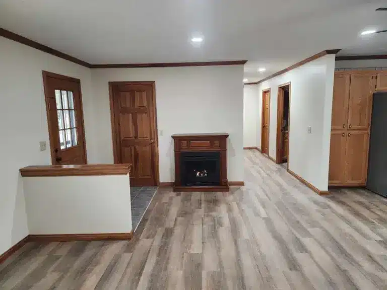 Open living room with gray vinyl plank flooring, wood-trimmed walls, a gas fireplace with oak surround, and stained wood doors.
