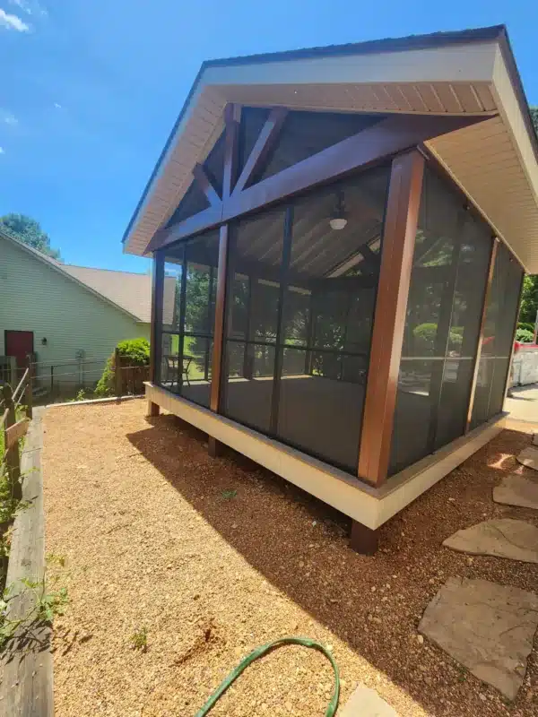 Freestanding screened porch with gable roof, dark wood posts, exposed beams, and ceiling fan on a gravel backyard site.