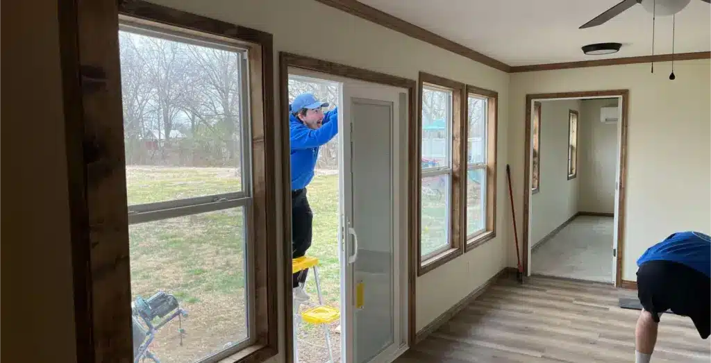 Worker on a step ladder installs a sliding glass door in a sunroom with wood-trimmed windows and vinyl plank flooring.
