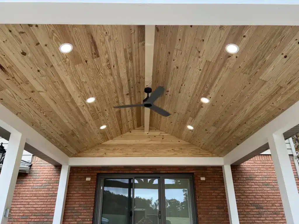 Vaulted tongue-and-groove pine ceiling on a covered porch with a black ceiling fan and recessed lights above a brick exterior.