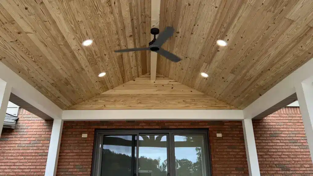 Vaulted tongue-and-groove pine ceiling on a covered back porch with a black ceiling fan, recessed lights, and brick walls.