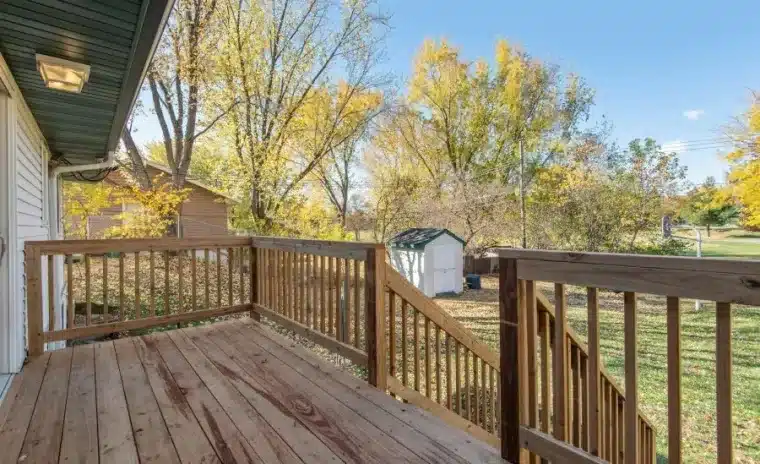 Wooden deck with spindle railing overlooking a grassy backyard with a small shed and autumn trees.